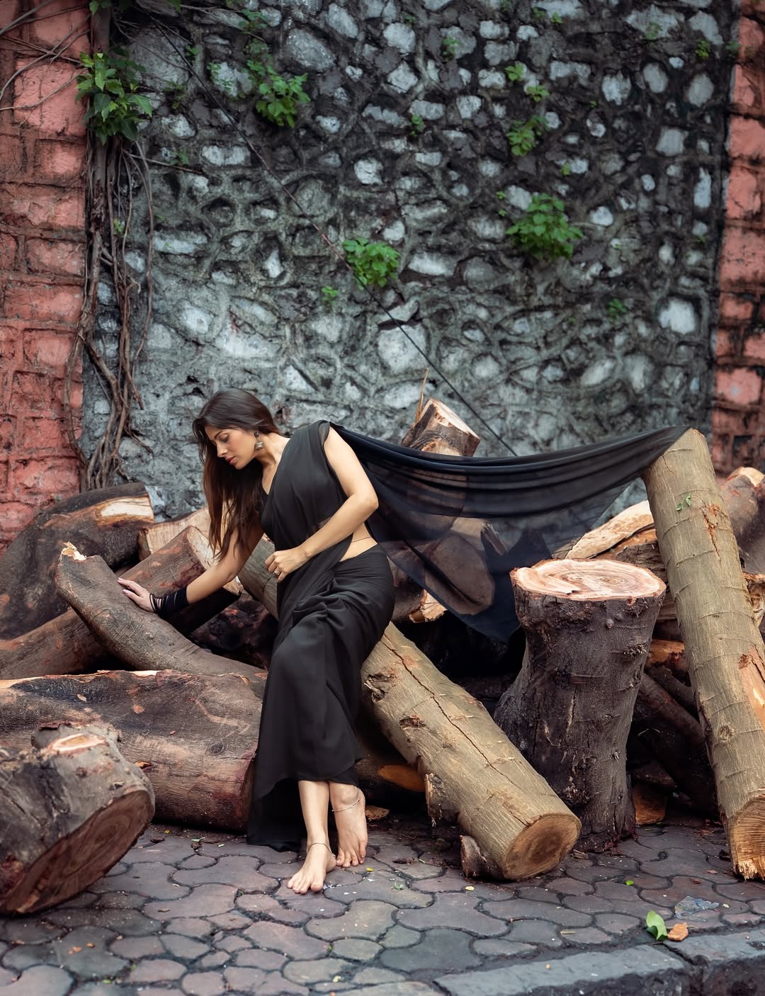 The rustic backdrop of logs contrasts with her elegant black saree.
